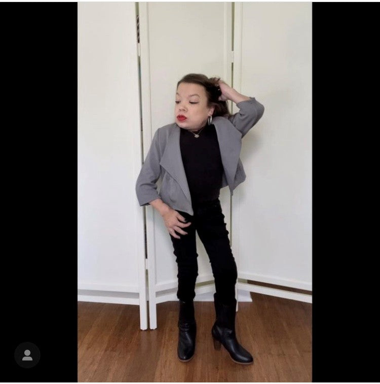 A little woman posing in an indoor setting wearing the jacket, black turtleneck, black pants, and black ankle boots.