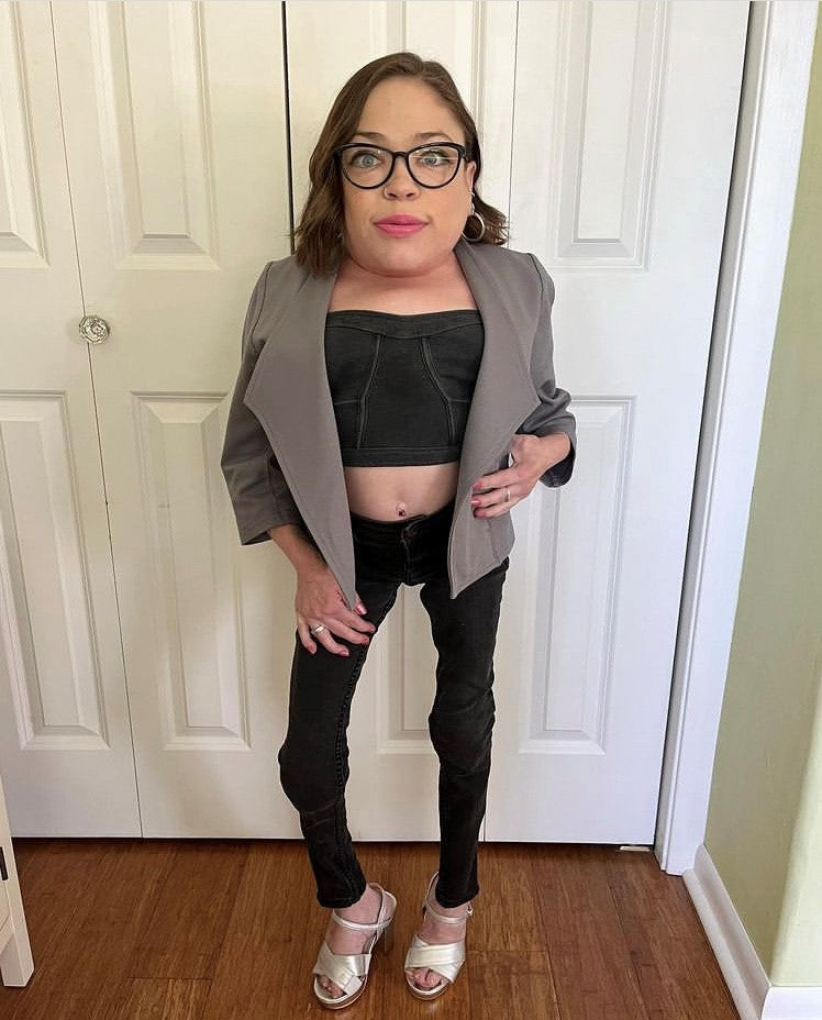 A little woman wearing the gray jacket, black bandeau top, black skinny jeans, and white heeled sandals stands in front of white closet doors.