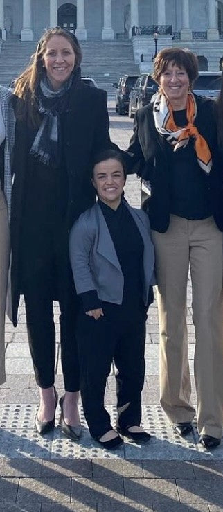 Model with dwarfism wearing the grey open-front jacket outdoors in a street setting, styled with black pants