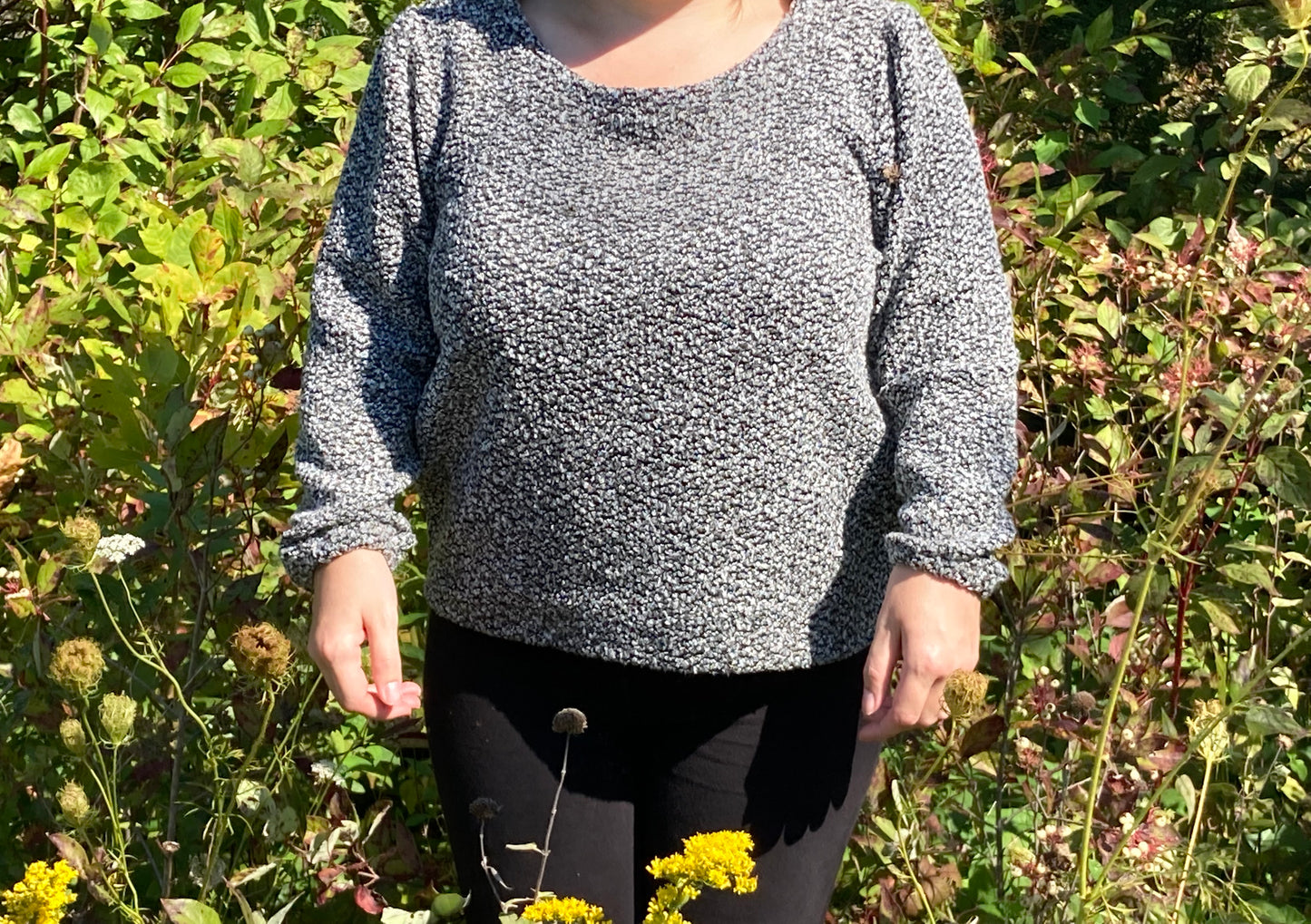 A little woamn wearing a grey knit sweater and black pants, standing outdoors in a garden with green foliage and yellow flowers.