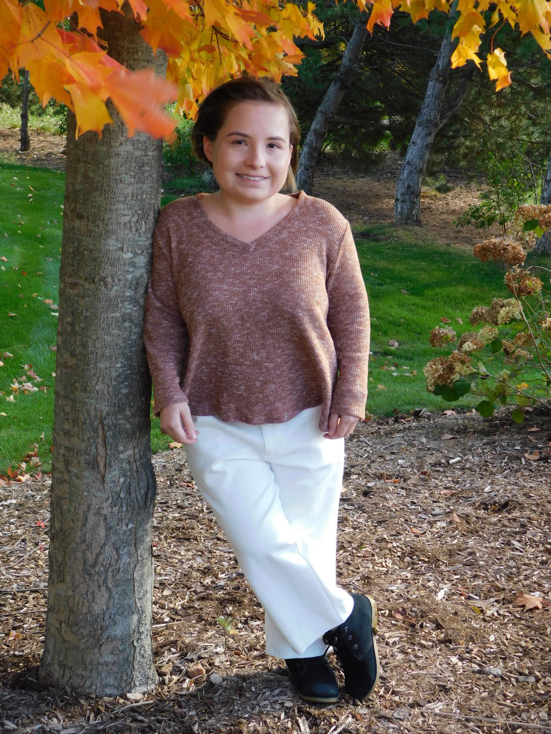 Lisa, founder of It Actually Fits, leaning against a tree in autumn, wearing a rose knit V-neck top and white wide-leg pants